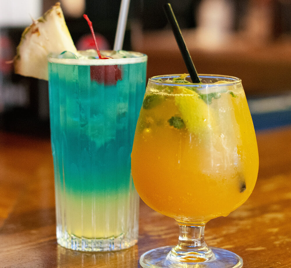 A blue cocktail with a pineapple slice and a cherry sitting next to a yellow cocktail