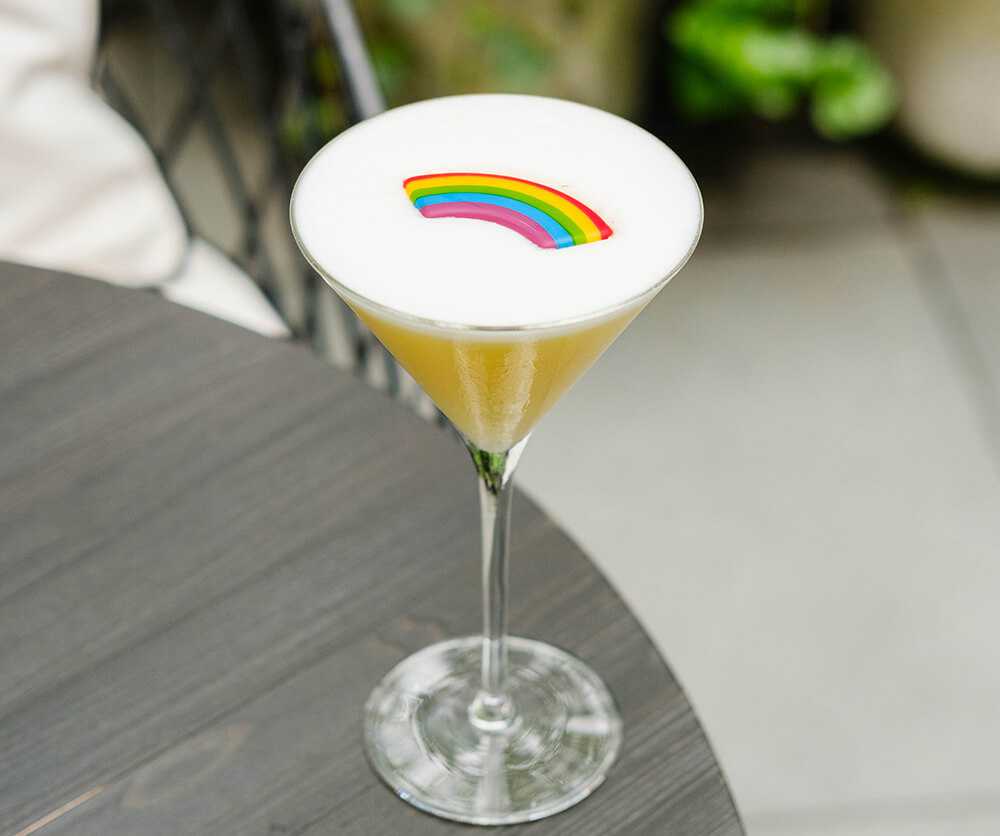 A yellow cocktail in a martini glass, with a candy rainbow sitting in the white foam on top