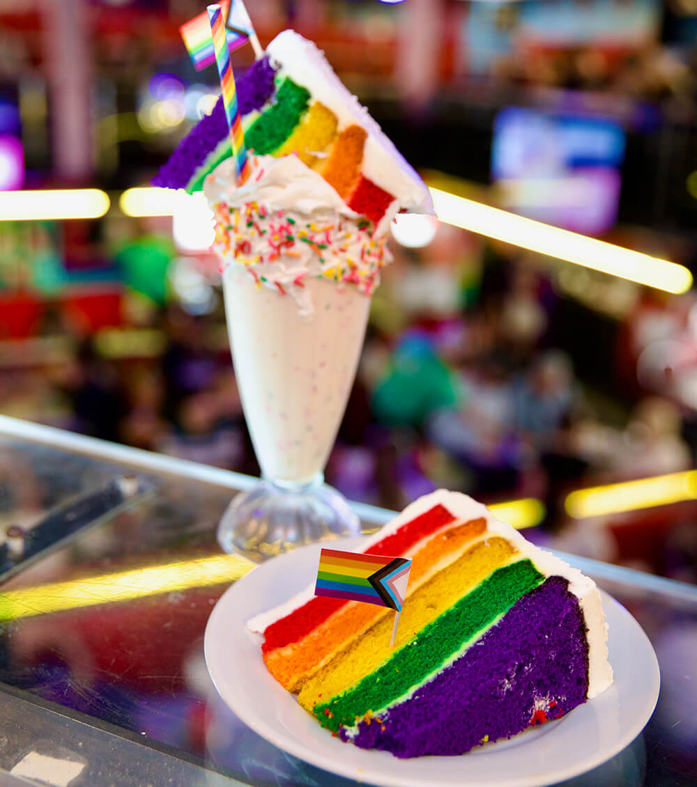 A slice of rainbow cake on a plate next to a milkshake topped with rainbow sprinkles, a rainbow straw, a whole slice of rainbow cake, and the inclusive pride flag on a toothpick