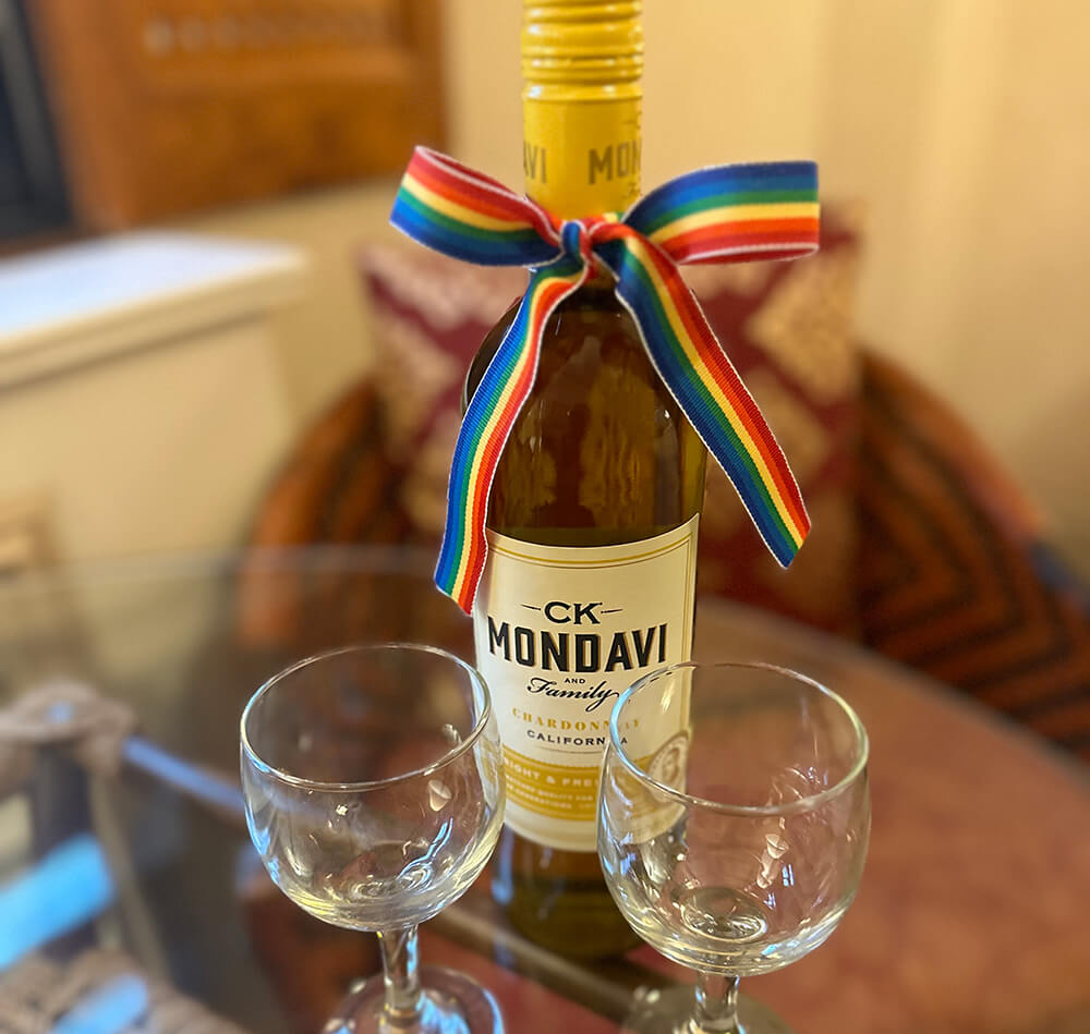 A bottle of CK Mondavi Chardonnay with a rainbow ribbon tied around the neck next to two wine glasses