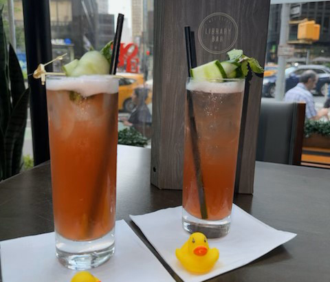 Two reddish-pink cocktails in tall glasses topped with cucumber slices with yellow rubber ducks sitting in front of the cocktails