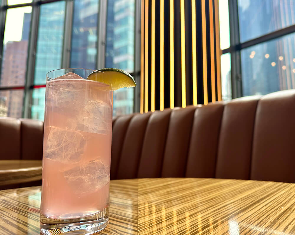 A light pink drink in a tall glass with a lime slice sitting on a table with a view of Times Square out the window