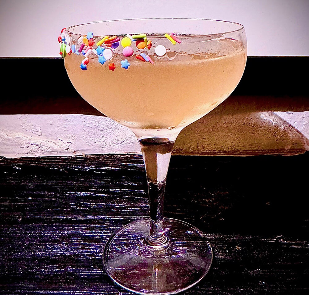 A peachy cocktail in a coupe glass with sprinkles on the rim