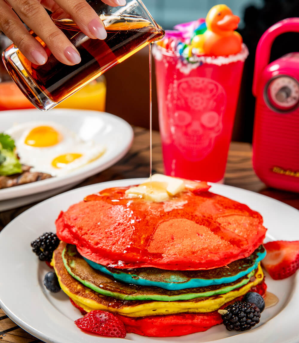 A hand pouring syrup over a rainbow stack of pancakes, with a bright magenta cocktail topped by rainbow decor in the background