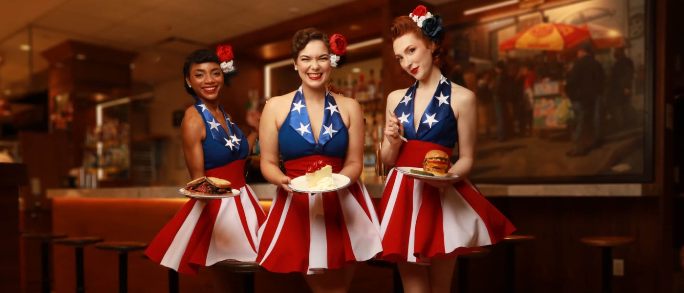 Three women in American-flag themed 1950s-style pinup attire, carrying plates with sandwiches and cheesecake