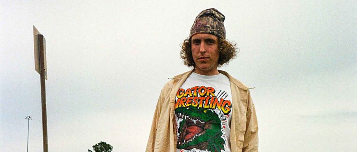 Andrew Callaghan, a white man with curly hair in a beanie and gator wrestling shirt, standing in grass by a highway