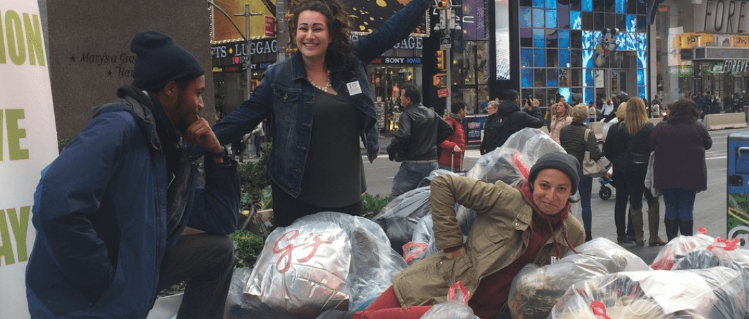 Three people standing amidst a mountain of bagged clothes at a Broadway Green Alliance textile recycling drive