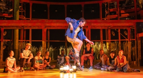 Ricky Ubeda dancing in front of the rest of the Broadway company of Illinoise, who are seated on the ground around a collection of lanterns